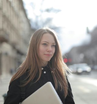 Confident young woman holding a laptop while walking on a bustling city street.